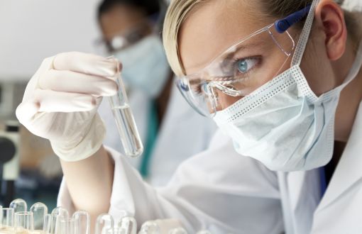 A female scientist in a white lab coat, lab goggles and mask is looking at and holding a test tube with a transparent liquid.