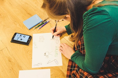 Dziewczyna w zielonym swetrze i spódnicy w kratę siedzi na podłodze i rysuje. Rysunek przedstawia kamerę, nuty, książkę oraz napis Tatry. Przed sobą ma telefon