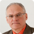 Zdjęcie portretowe: prof. Jan Awrejcewicz w brązowym garniturze i łososiowej koszuli na szarym tle.
