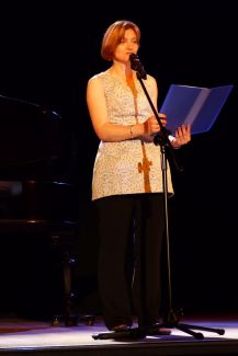 Kobieta (prof. M. Buczkowska) w białej bluzce i czarnych spodniach stoi na scenie przed mikrofonem. W rękach trzyma niebieską, rozłożoną teczkę.