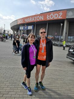 Prof. Wójcik z żoną, obydwoje w strojach sportowych przed dużą, zaokrągloną halą z dużym pomarańczowym banerem: DOZ Maraton Łódź.