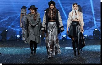 Cztery modelki w szaro czarnych strojach na wybiegu podczas pokazy mody Gala Politechnika Fashion. W tle niebieska, oświetlona ściana.