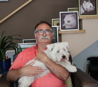 Prof. Tomasz Kapitaniak w łososiowej polówce siedzi w fotelu i trzyma na rękach białego psa. W tle ściana ze zdjęciami tego samego psa.