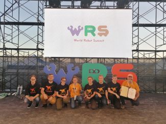 9 członków zespoły Raptors w czarnych koszulkach z logo zespołu. Nad nimi ekran z napisem WRS - World Robot Summit.