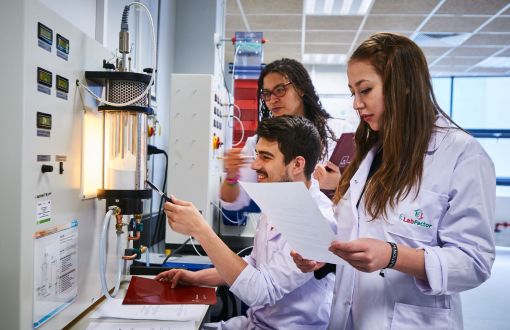 Three TUL students in lab coats are standing sideways by a white lab machine. The student sitting in the middle is showing something on it.