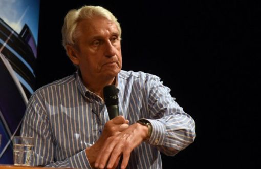 Bogusław Wołoszański w koszuli w paski trzyma w ręku mikrofon, drugą ręką gestykuluje.
