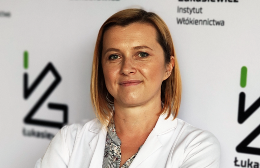 Zdjęcie portretowe: Magdalena Olak-Kucharczyk na tle ścianki z logo Sieć Badawcza Łukasiewicz.