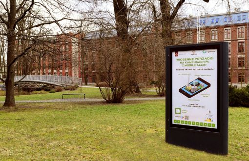 Kampus B Politechniki Łódzkiej: na pierwszym planie citylight z plakatem reklamowym. W tle park z niewielką ilością zieleni oraz Budynek Trzech Wydziałów.