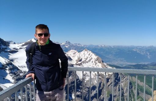 Paweł Czapski ubrany na sportowo na tarasie widokowym wysoko w górach. W tle ośnieżone góry i bezchmurne niebo.