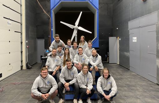 14 studentów w szarych bluzach, członków Zespołu GUST z Politechniki Łódzkiej, siedzą w trzech rzędach. W tle ich turbina.