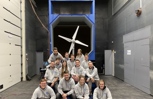 14 studentów w szarych bluzach, członków Zespołu GUST z Politechniki Łódzkiej, siedzą w trzech rzędach. W tle ich turbina.