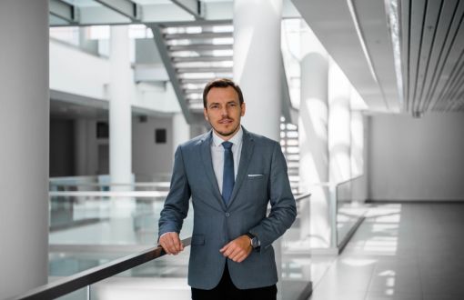 dr Dariusz Koperczak w szarym garniturze stoi na tle przestrzeni w nowoczesnym gmachu