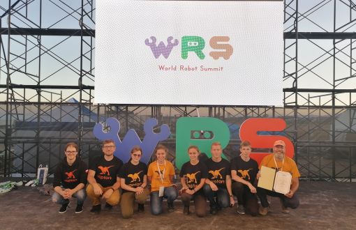 9 członków zespoły Raptors w czarnych koszulkach z logo zespołu. Nad nimi ekran z napisem WRS - World Robot Summit.